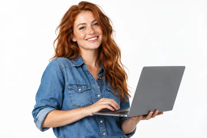 Eine freundlich wirkende rothaarige Marketingfachkraft in einem Jeanshemd hält einen aufgeklappten Laptop vor einem weißen Studiohintergrund.